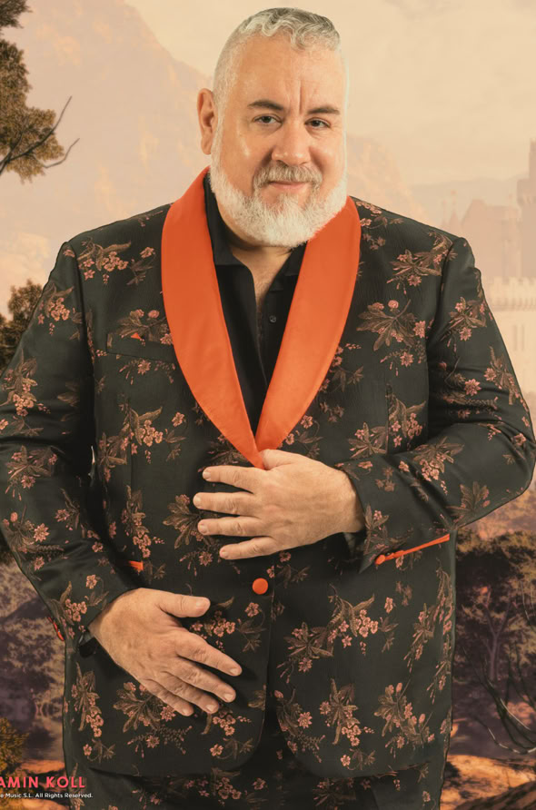 A man with a beard wearing a floral-patterned suit with an orange collar, standing in front of a scenic background.