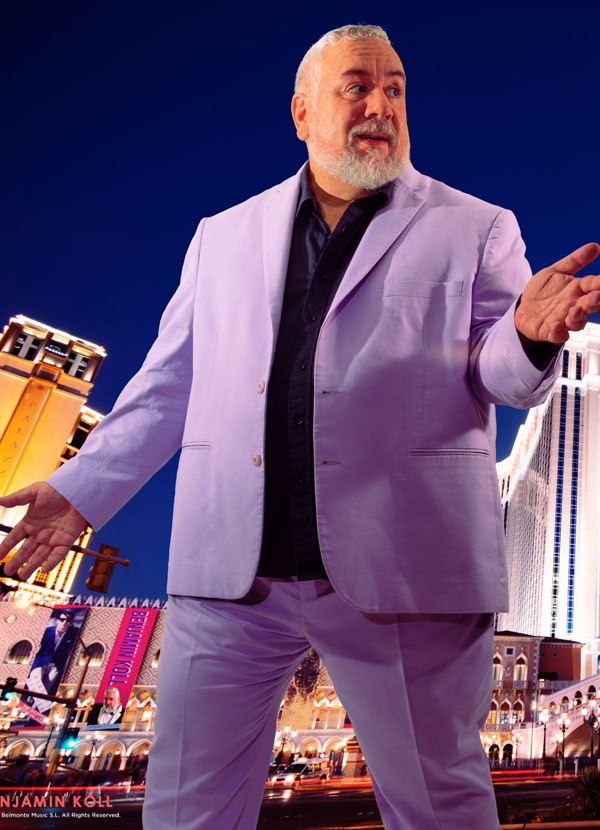 A man with a beard wearing a light purple suit gestures with one hand, standing in front of a cityscape with brightly lit buildings at night.