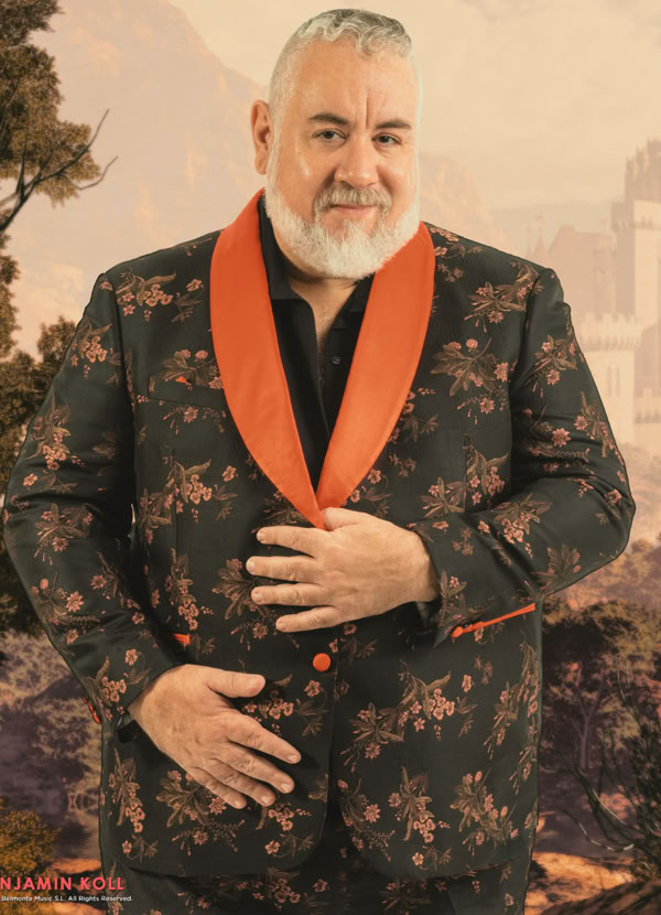 A man with a beard wearing a floral-patterned suit with an orange collar, standing in front of a scenic background.