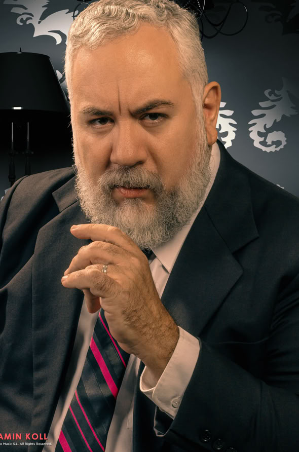A man with a gray beard wearing a suit and tie, sitting in front of a patterned wallpaper with a lamp in the background.