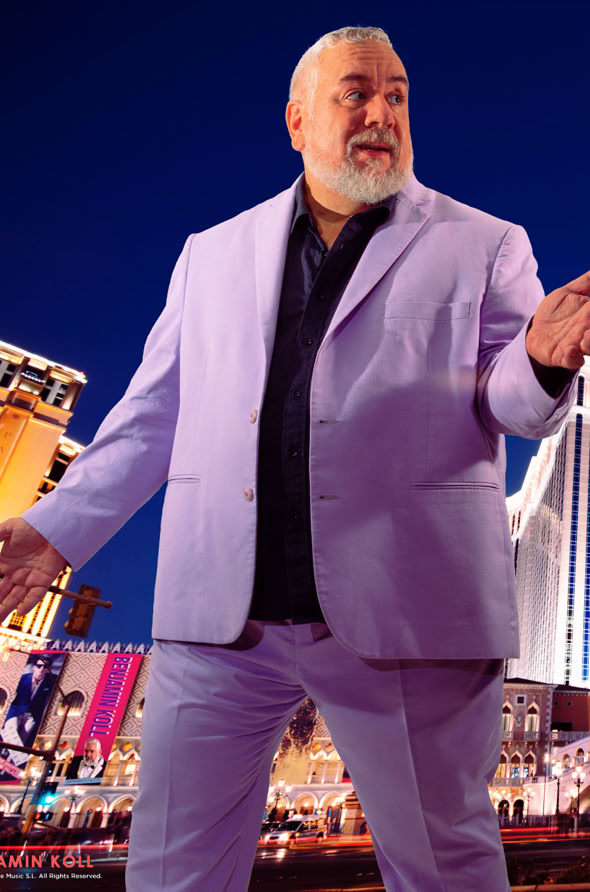 A man with a beard wearing a light purple suit gestures with one hand, standing in front of a cityscape with brightly lit buildings at night.