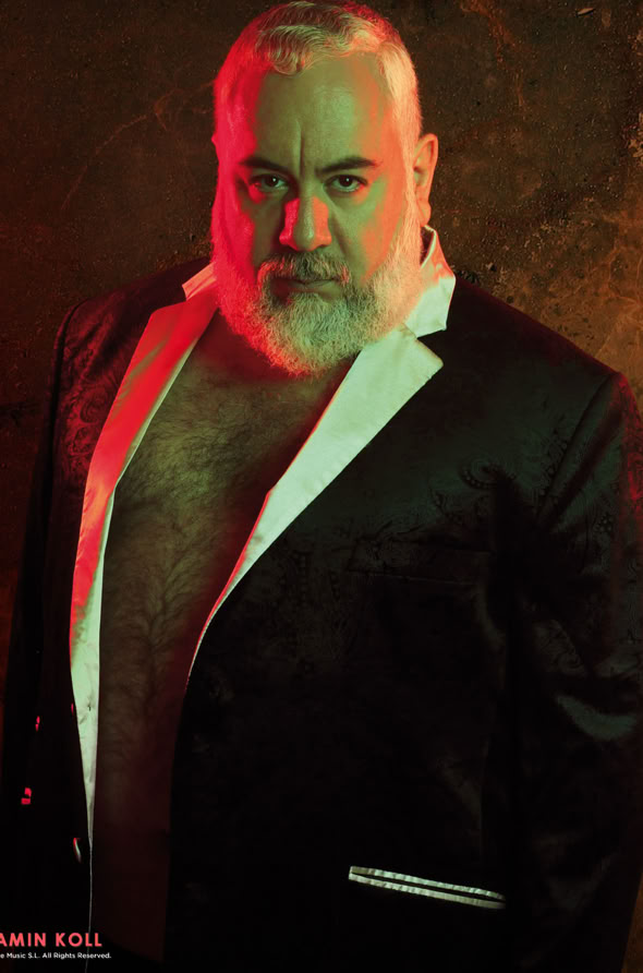 A man with a beard wearing a black suit jacket with white lapels, standing against a textured background with red and green lighting.