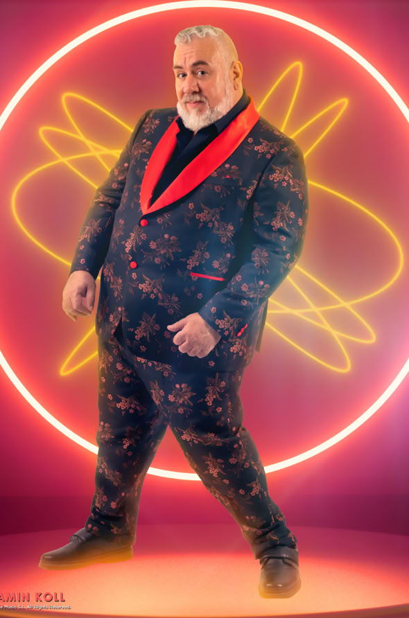 Man in a patterned suit with a red collar stands in front of a neon circular design on a pink background.
