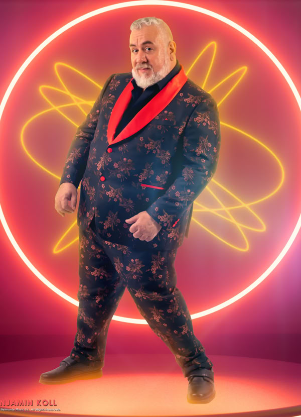 Man in a patterned suit with a red collar stands in front of a neon circular design on a pink background.