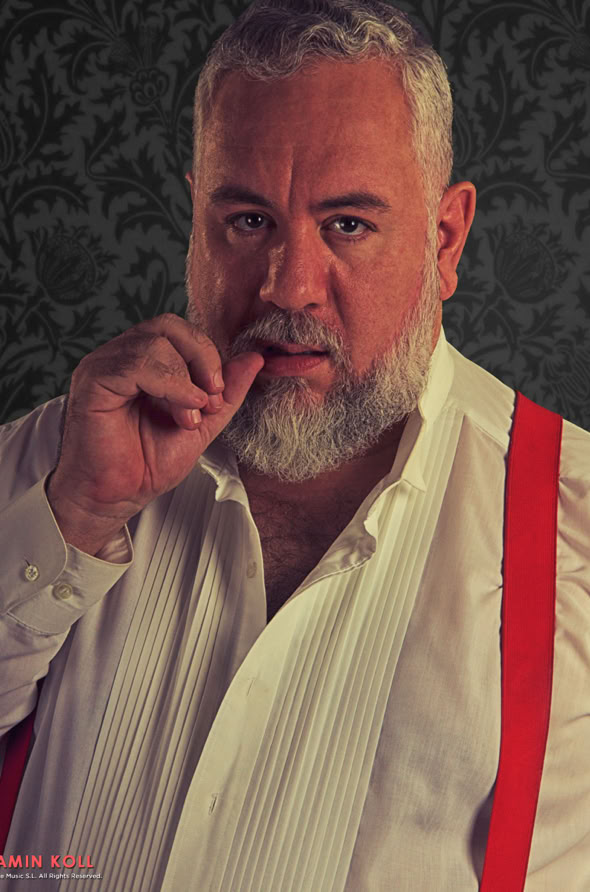 A man with a beard and gray hair wearing a white shirt and red suspenders, standing against a dark patterned background, touching his lips with his finger.